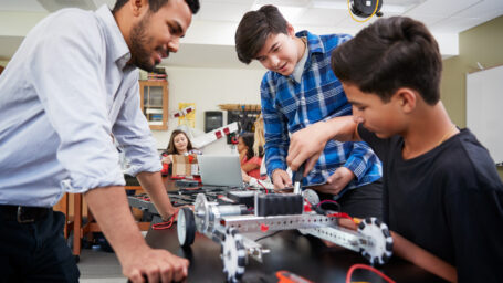 A male teacher watches two young male students as they build a robotic car on a table.