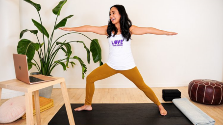 Woman with long dark hair and a white tank top doing a warrior yoga pose on a black yoga mat