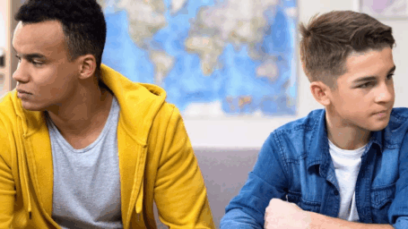 Two teenage boys sitting apart looking upset after an argument in a classroom with world map on wall