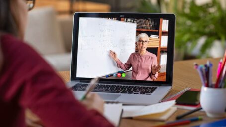 Young student watching lesson online and studying from home. Young woman taking notes while looking at computer screen following professor doing math on video call. Latin girl student studying from home and watching teacher explaining math formula on video chat.