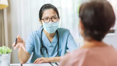 Asian Woman in blue uniform and protective face mask giving advice to Senior female patient at hospital. Practitioner wear eyeglasses and surgical mask writing information on prescription