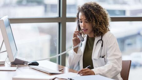The mid adult female doctor, sitting in her office, uses the landline to make phone calls regarding patient records.